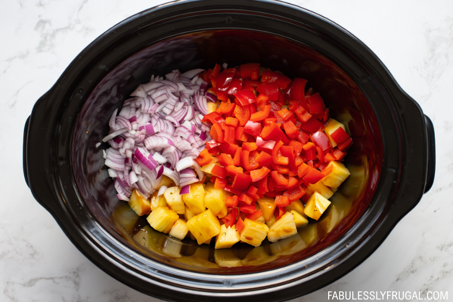 Raw chicken, pineapple, onion, and bell peppers placed into slow cooker – fabulesslyfrugal.com branding
