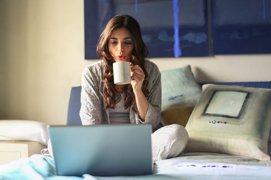 Woman sipping coffee and working on her laptop sitting on bed